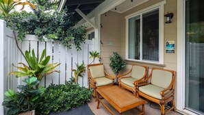 A covered patio with a wooden table and four wicker chairs, surrounded by lush greenery and a white fence. - Modern Family Beach Villa near shops and dining (Kapolei)