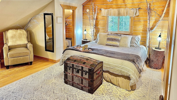 A cozy bedroom with a sloped ceiling, a large bed with multiple pillows, a chair, a small table, and a trunk. - Waterfront Log Cabin on Airstrip with Aurora Views (Fairbanks)