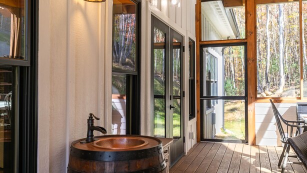 Eine Holzterrasse mit einem rustikalen Fassbecken, großen Glasüren, die in einen bewaldeten Bereich führen, und einem Esstisch mit Stühlen.