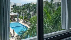 Blick auf einen Swimmingpool mit Liegestühlen und Palmen, sichtbar durch ein Fenster mit Jalousien.