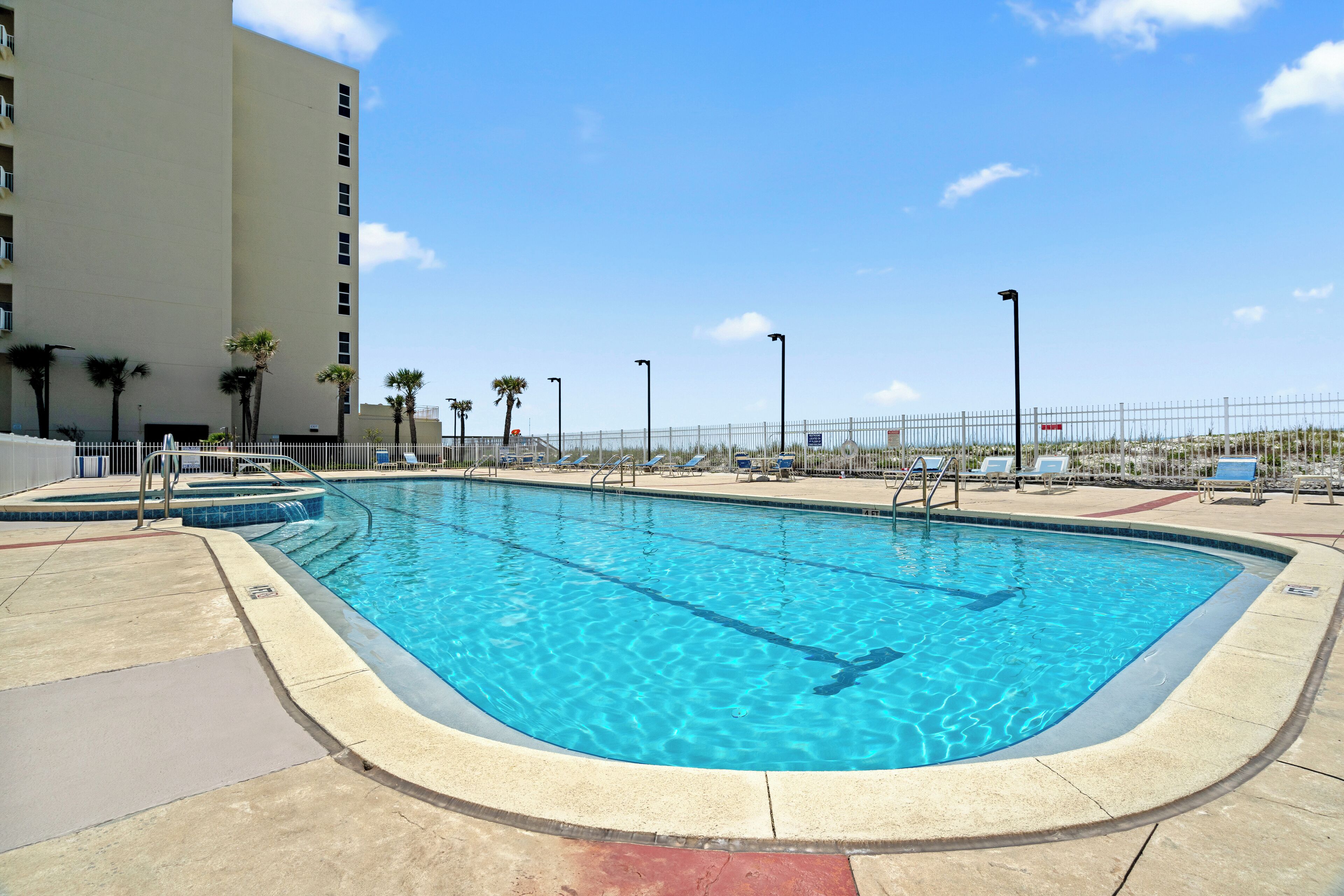 A rectangular swimming pool with clear blue water, surrounded by a white fence, and adjacent to a multi-story building.