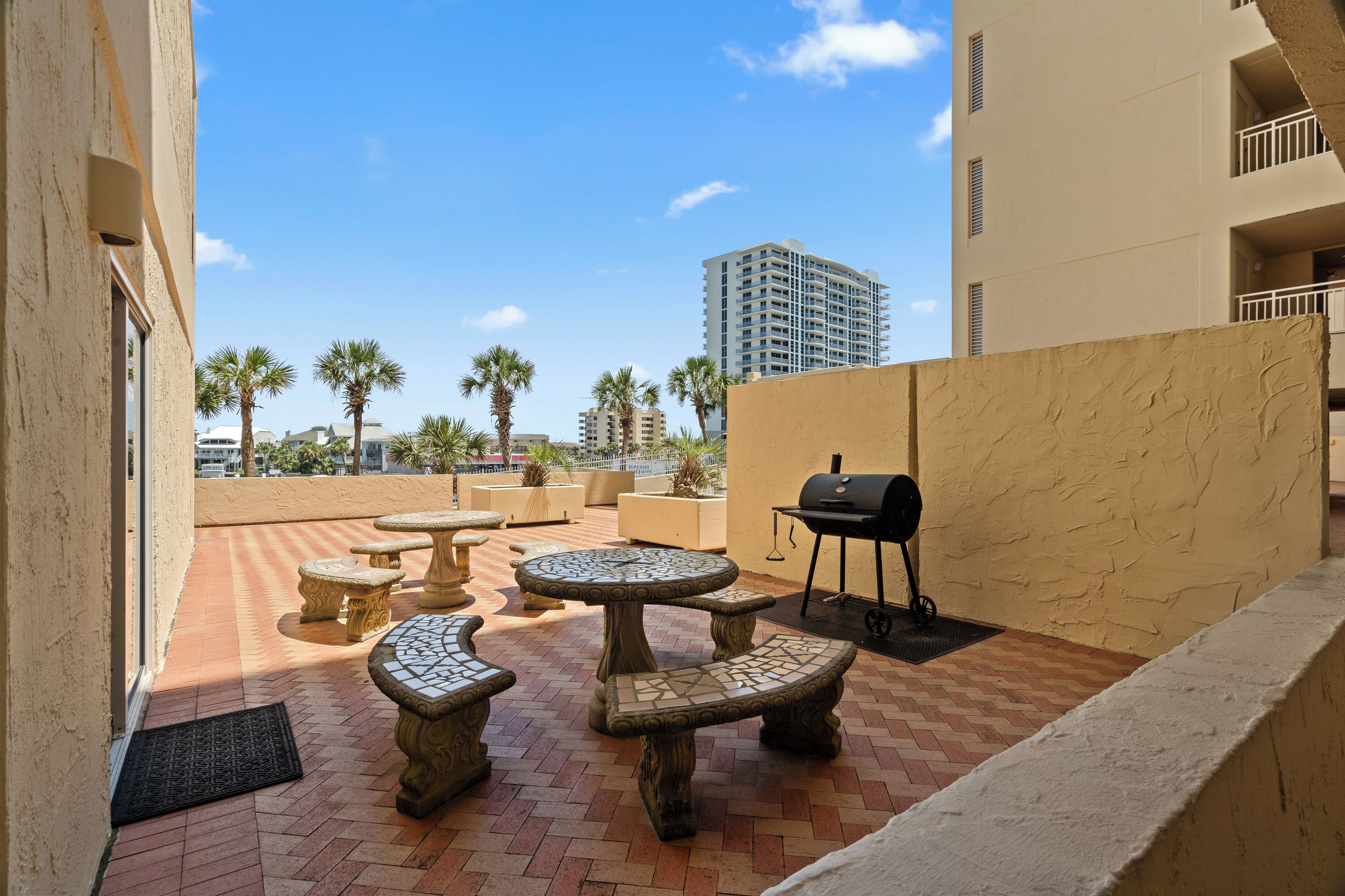Un patio al aire libre con mesas y bancos de piedra, una parrilla y vistas a palmeras y edificios al fondo.