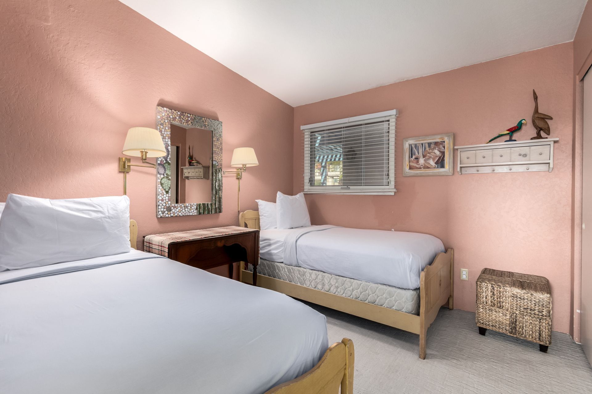 A bedroom with two beds, a desk, a chair, a small table, a window with blinds, a mirror, and a picture on the wall.