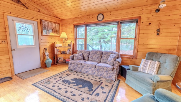 Un interior de cabaña acogedor con techo de madera, un sofá, una silla, un tapete con estampado de oso y un ventanal con vistas a árboles.