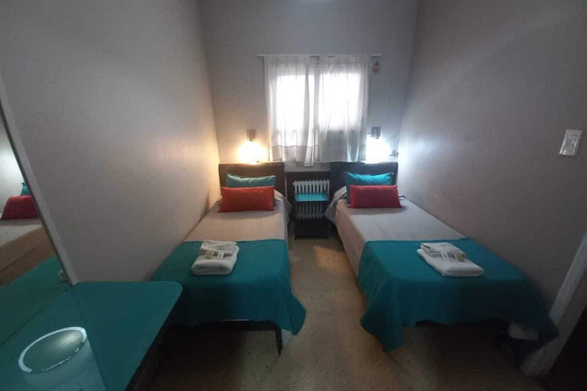 A small, compact hotel room with two single beds, each with a teal bedspread and red pillows. There is a small table with a glass of water and a folded towel on it. A radiator is visible in the background, and a window with curtains allows natural light into the room.