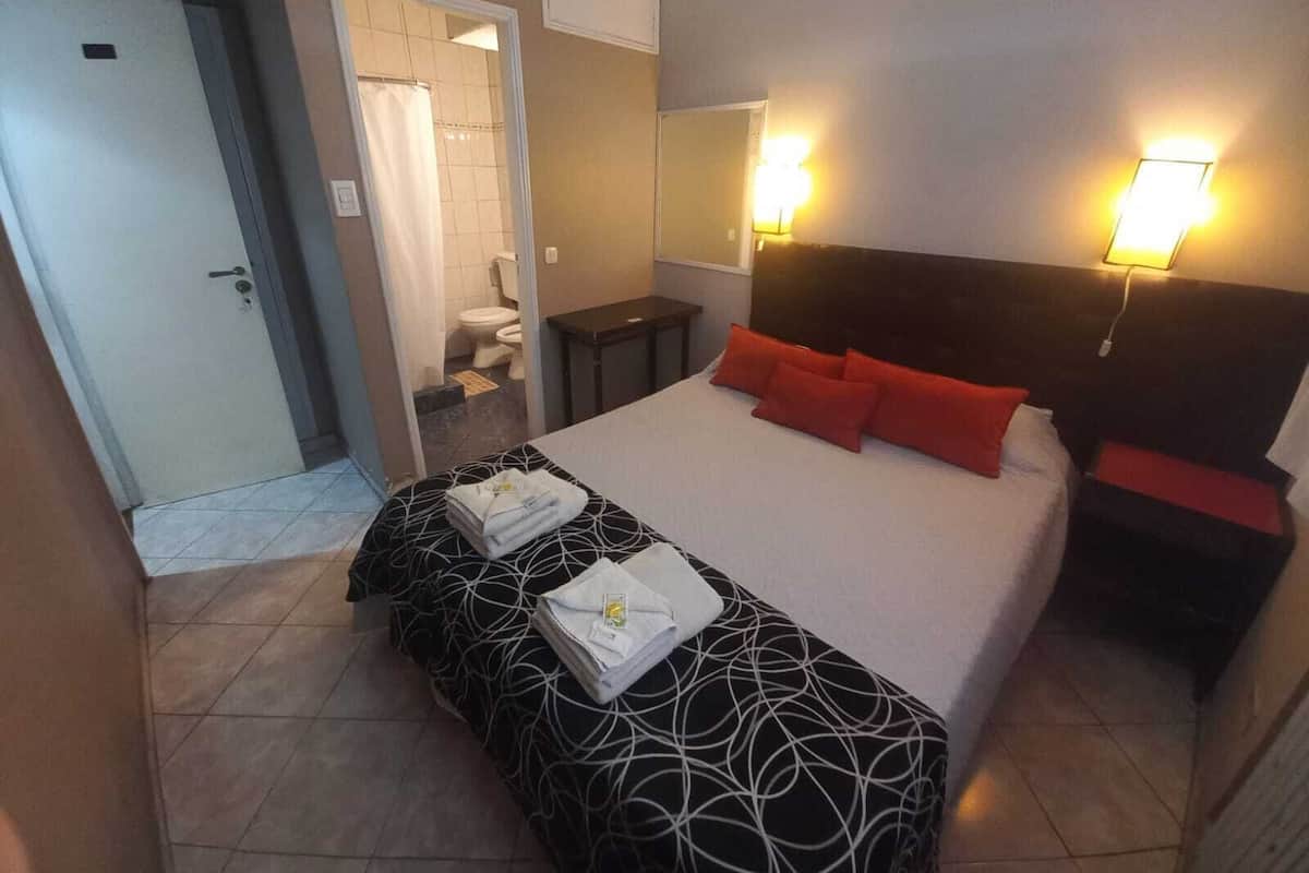A hotel room with a bed, a bedside table, a mirror, and a bathroom visible through an open door.