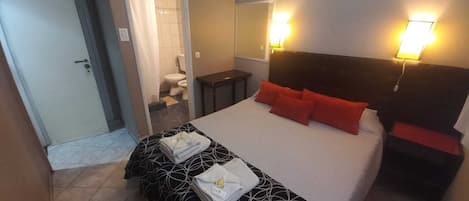Quarto de hotel com cama, mesa de cabeceira, espelho e banheiro visível através de uma porta aberta.