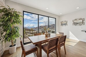 Un comedor con una mesa grande y sillas, una planta en maceta y vistas de montañas a través de amplios ventanales.