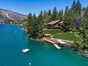 A lakeside property with a wooden dock, a covered seating area, and a large house with a balcony overlooking the water.