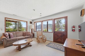 A living room with a sofa, coffee table, wicker chair, and large windows with a view of mountains.