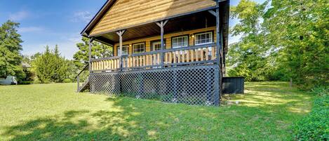 A wooden cabin with a porch, surrounded by a grassy yard and trees.