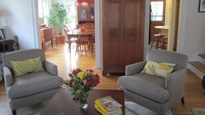 A living room with two armchairs, a coffee table with books and a vase of flowers, a wooden cabinet, and a dining area in the background. - Classic Ann Arbor Home Near Stadium and Campus (Ann Arbor)