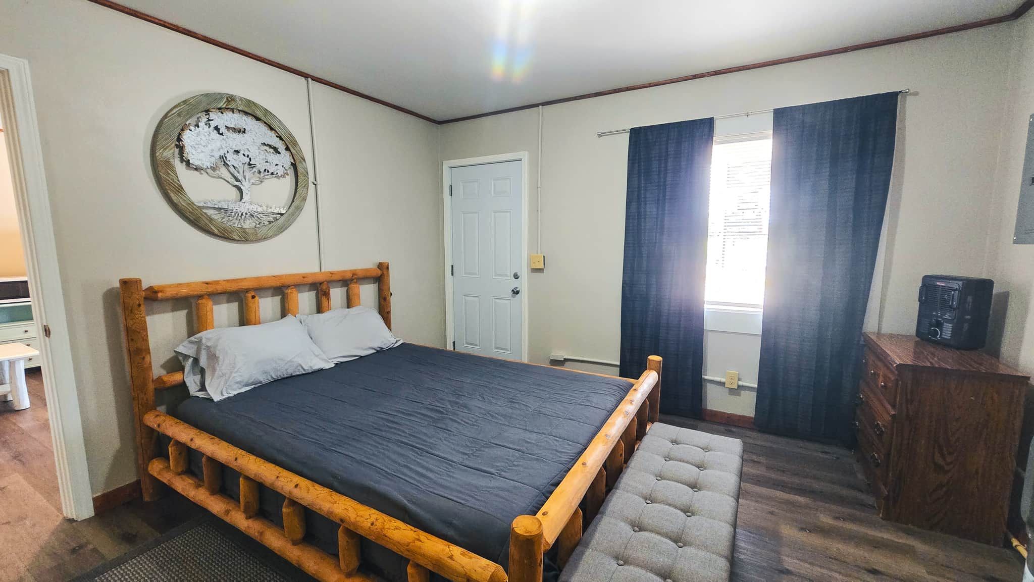 Un dormitorio con una cama de madera, una puerta, una ventana con cortinas y un pequeño armario de madera.