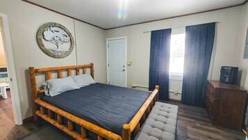 A bedroom with a wooden bed, a door, a window with curtains, and a small wooden cabinet.