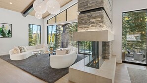 Un salon moderne avec une cheminée en pierre, des canapés blancs et de grandes fenêtres donnant sur la forêt.