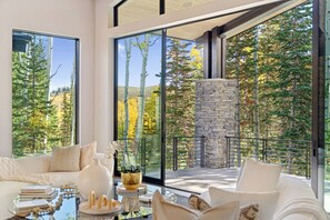 A modern living room with a large glass wall, a stone fireplace, and a view of autumn trees. - Ski-In/Ski-Out Alpine Estate with Pool, Hot Tub, Climbing Wall! (Park City)