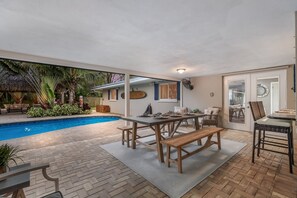 A poolside area with a dining table and chairs, a bench, and a small table with a fan. - Tropical Tiki:Outdoor Oasis, Tiki Hut, Pool by Anna Maria Life Vacation Rentals (Bradenton)