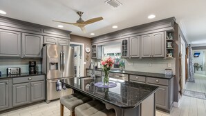 A modern kitchen with grey cabinets, stainless steel appliances, a central island with bar stools, and a ceiling fan. - Tropical Tiki:Outdoor Oasis, Tiki Hut, Pool by Anna Maria Life Vacation Rentals (Bradenton)