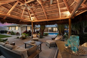 An outdoor patio with a thatched roof, wicker furniture, and a pool in the background. - Tropical Tiki:Outdoor Oasis, Tiki Hut, Pool by Anna Maria Life Vacation Rentals (Bradenton)