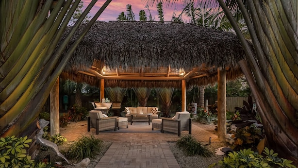 A covered outdoor seating area with wicker furniture and a thatched roof, surrounded by tropical plants and a brick pathway. - Tropical Tiki:Outdoor Oasis, Tiki Hut, Pool by Anna Maria Life Vacation Rentals (Bradenton)