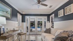 A room with a white sofa, a dining table, and a ceiling fan. There is a view of a pool outside through a glass door. - Tropical Tiki:Outdoor Oasis, Tiki Hut, Pool by Anna Maria Life Vacation Rentals (Bradenton)