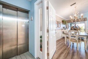 A hallway with a double door, a dining area with a chandelier, and a kitchen visible through an open door. - Palm Gables 112 :NO ELEVATOR Jul 25 - Nov 15, 2026 - Gulf Front Condo, Pool, Beach Access (Holmes Beach)