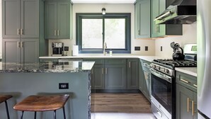 Una cocina con armarios verde oscuro, un ventanal grande, una estufa y dos taburetes de bar.