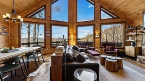 A spacious wooden cabin with large windows, a dining area, a living room with a sofa and chairs, and a stone fireplace. - Grandeur View Lodge - Mountain View | Hot Tub | Game Room (Blue Ridge)