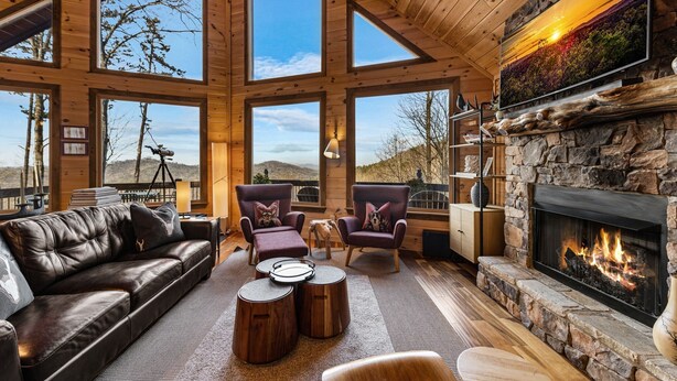 A spacious room with a stone fireplace, leather sofa, and large windows offering a view of the outdoors. - Grandeur View Lodge - Mountain View | Hot Tub | Game Room (Blue Ridge)