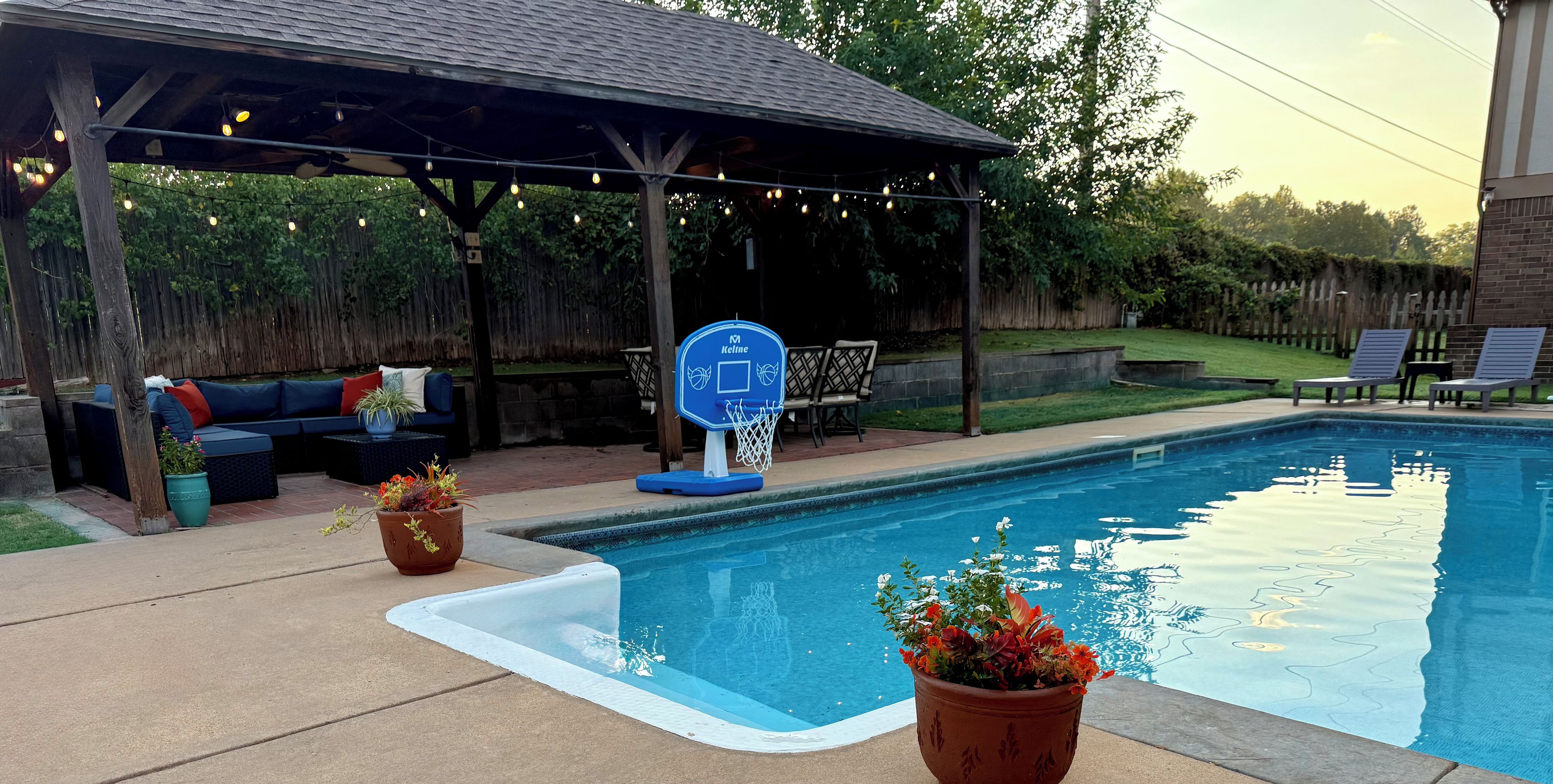 A backyard with a swimming pool, a covered seating area, and outdoor furniture.