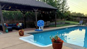 A backyard with a swimming pool, a covered seating area, and outdoor furniture.