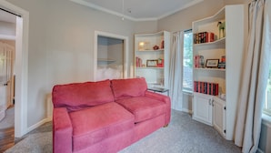 Ein Wohnzimmer mit einem roten Sofa, einem Deckenventilator, einem Bücherregal und einem fensterbelaubten Fenster.