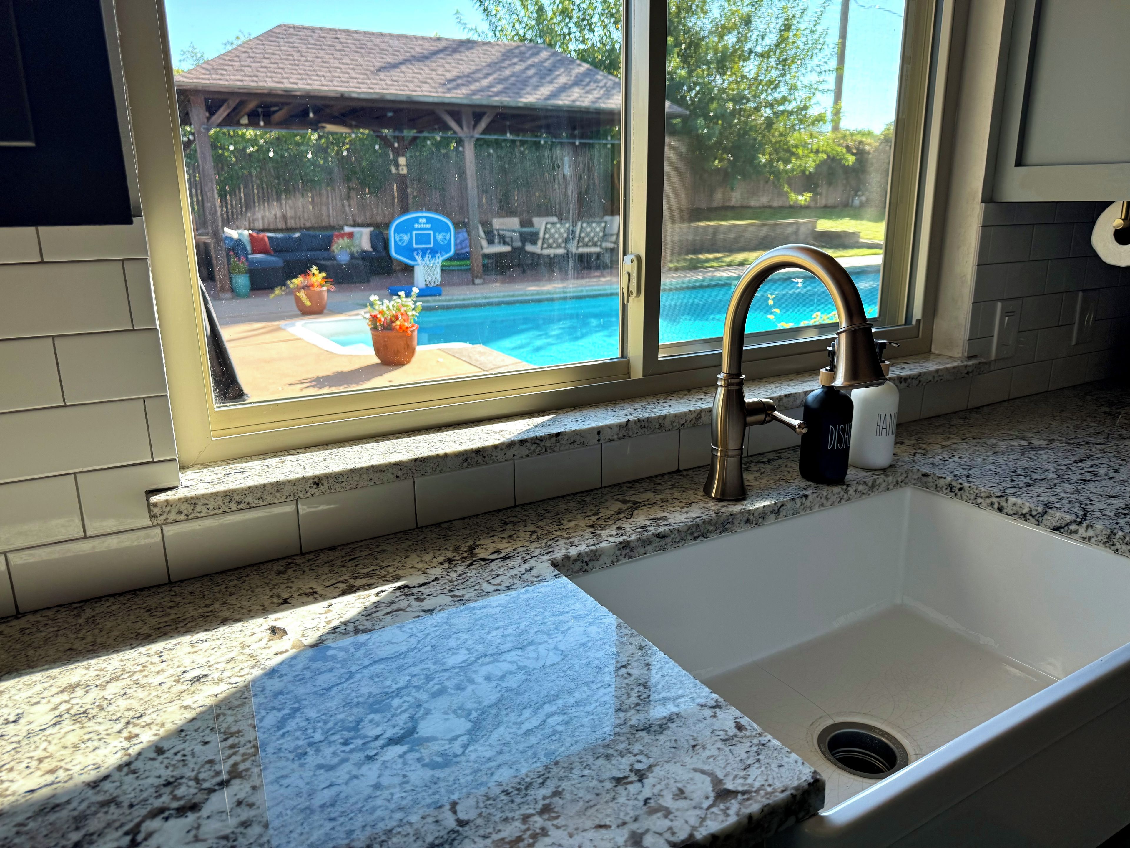 A kitchen with a granite countertop, a stainless steel sink, and a window overlooking a backyard with a pool and outdoor furniture.