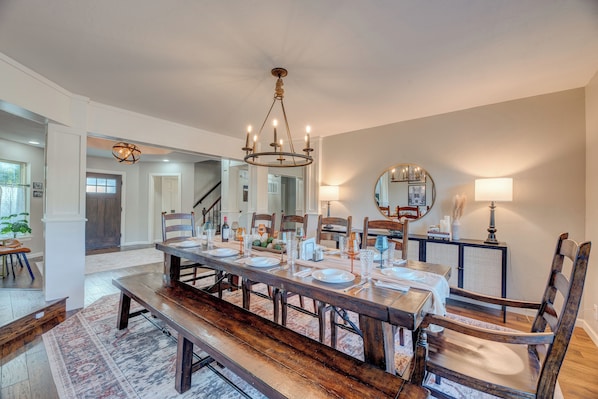 A dining area with a long wooden table set for six, surrounded by wooden chairs. A chandelier hangs from the ceiling, and a mirror is mounted on the wall above the table. - Heated Pool, 3 King Bedrooms- 2 Suites, Sleeps 14, Private Backyard Near OU (Norman)