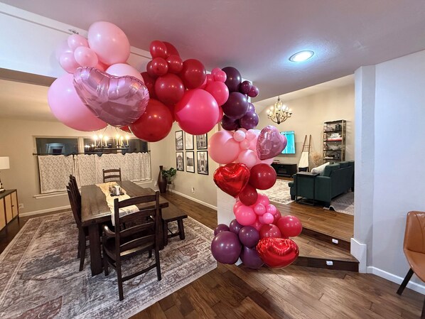 A dining area with a wooden table and chairs, a chandelier, and a living room in the background. - Heated Pool, 3 King Bedrooms- 2 Suites, Sleeps 14, Private Backyard Near OU (Norman)