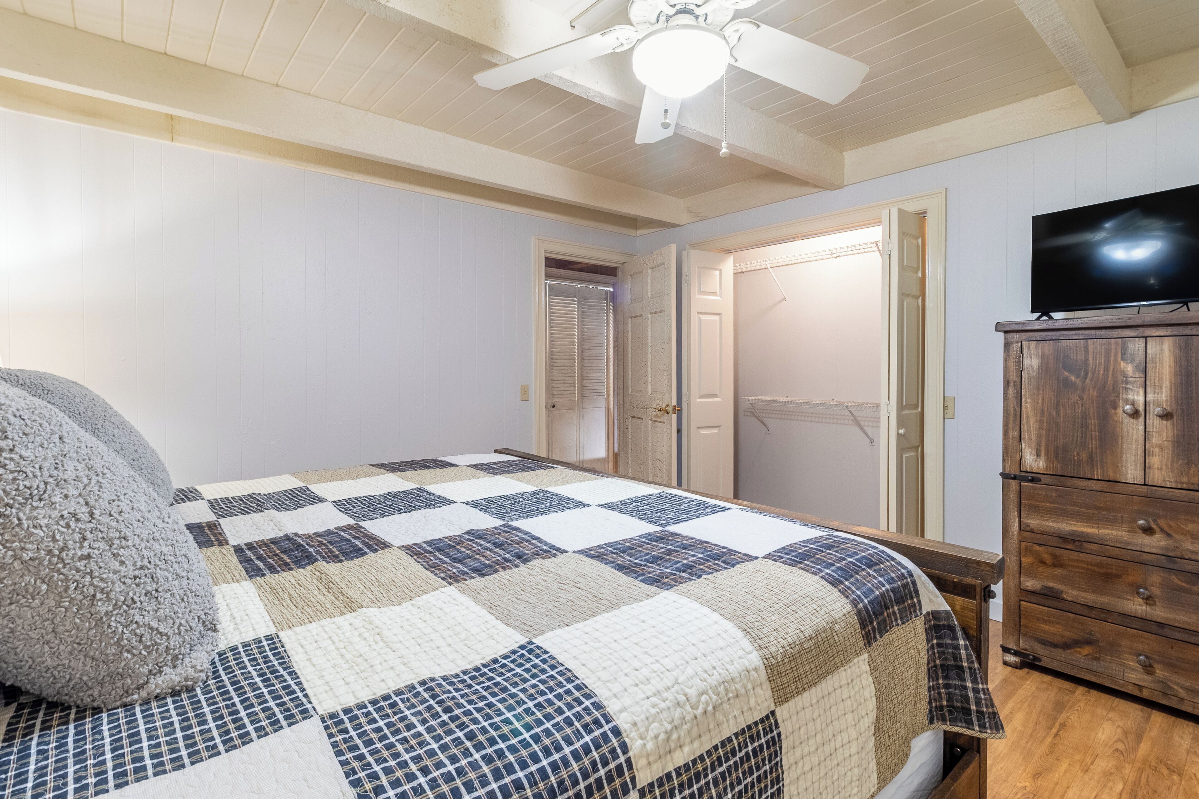 A bedroom with a checkered quilt, a wooden dresser, and a ceiling fan.