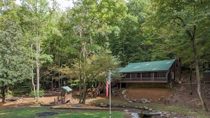 A cabin with a green roof, surrounded by trees and a small stream.