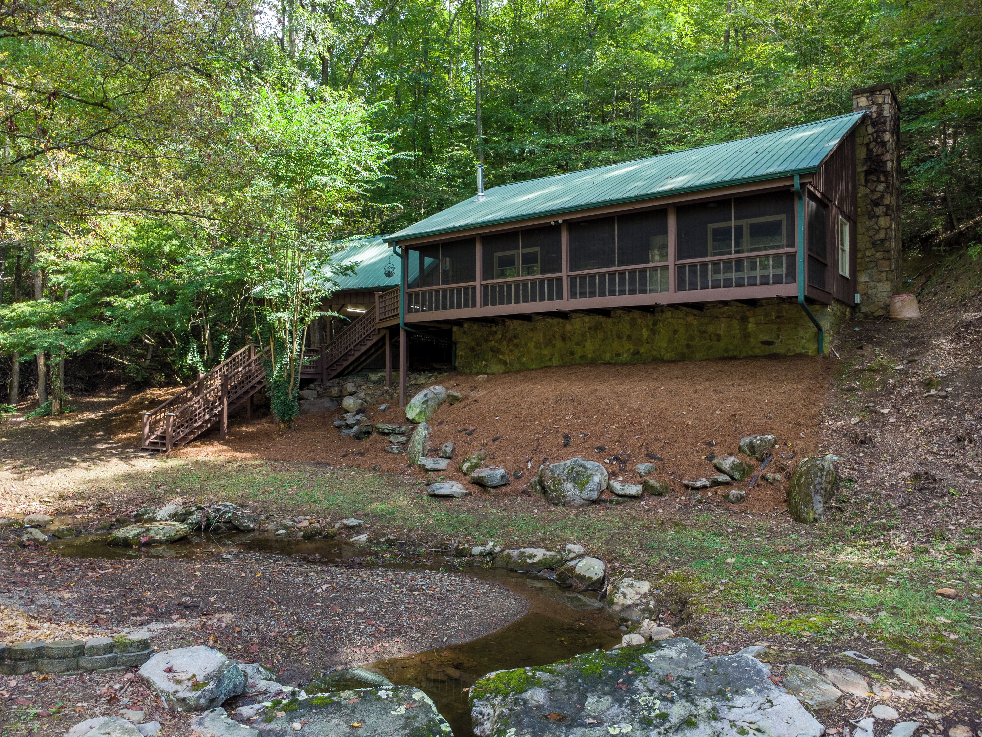 Eine Holzhütte mit Steinunterbau und grüner Dachdeckung, umgeben von bewaldeter Landschaft mit einem kleinen Bach und einer Holztreppe, die zu ihr hinaufführt.