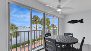 A balcony with a table and chairs, a ceiling fan, and a view of a body of water with palm trees. - Bayfront Bliss Near Vanderbilt Beach (Naples)