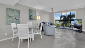 Ampla sala de estar com mesa de jantar, cadeiras brancas, sofá cinza e varanda com vista para palmeiras.