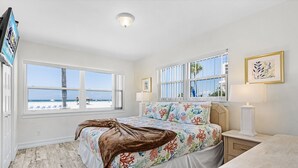 A bedroom with a large window, a bed with a floral bedspread, a bedside table with a lamp, and a view of the beach. - Combined Beachfront Bliss - Aloha Kai #24 & #24N - Roelens (Siesta Key)