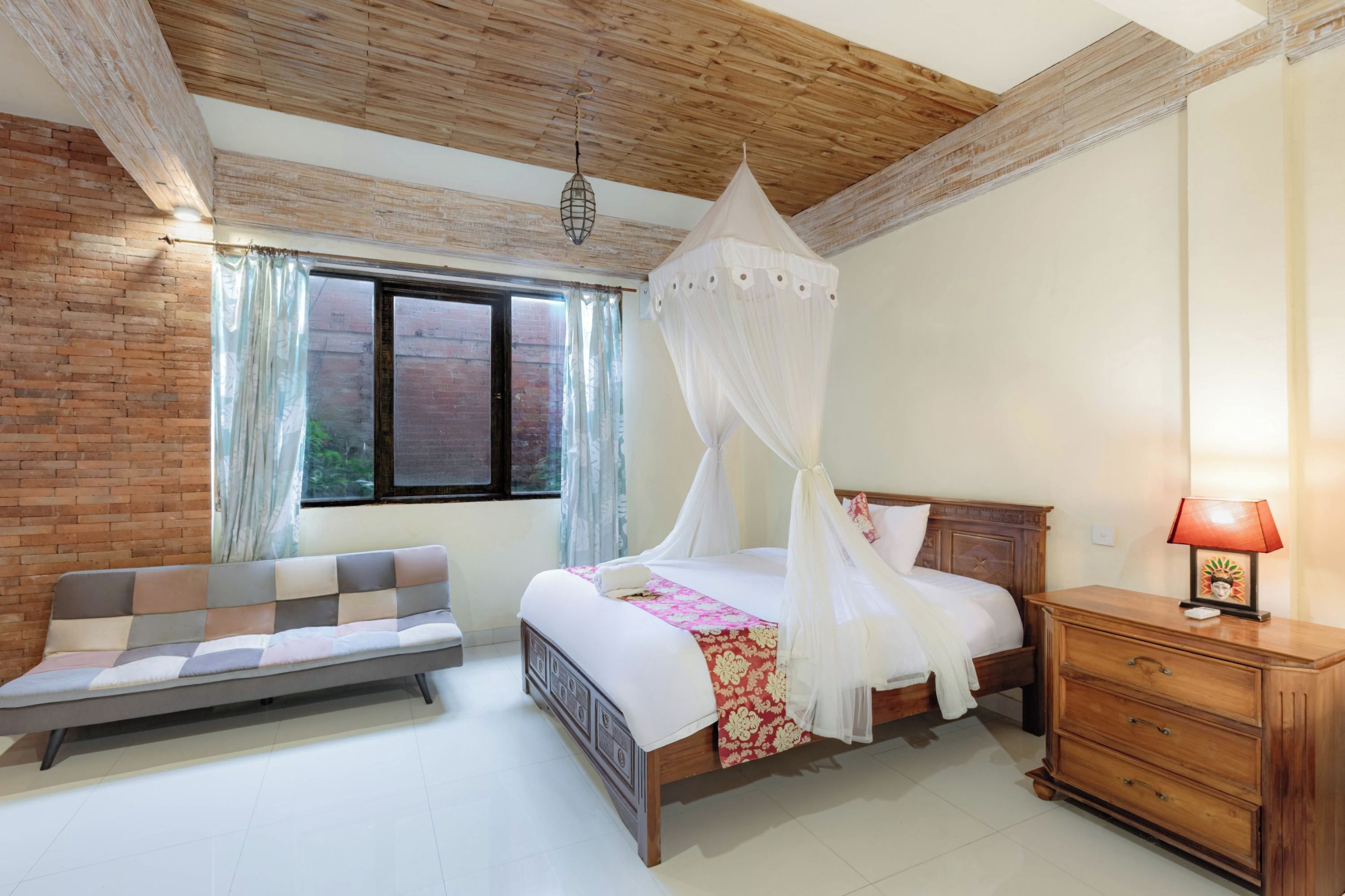 Quarto com cama, mesa de cabeceira, um banco de madeira e janela com cortinas.