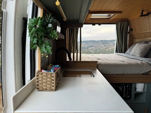 Un interior acogedor con una cama, una mesita, una cesta y una ventana con vistas a un paisaje pintoresco.