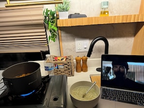 Una cocina con estufa a gas, una olla encima, un tazón de fideos, una computadora portátil y algunos condimentos.