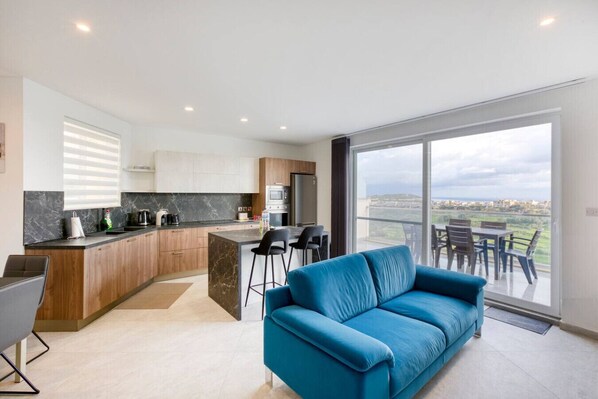 A modern kitchen with a marble countertop, wooden cabinets, and a built-in oven. There is a blue sofa in the living area with a view of the city through large windows.