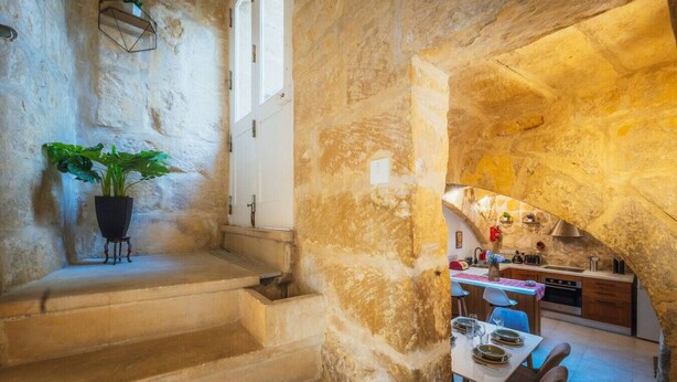 Una escalera de piedra conduce a un comedor con una mesa lista para dos comensales, una planta en maceta y una cocina visible a través de un arco.