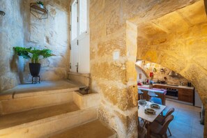 Un escalier en pierre mène à une salle à manger où une table est dressée pour deux personnes. On y aperçoit également une plante en pot et, à travers une arche, une cuisine.