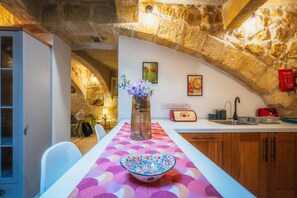 Eine Küche mit Steinwänden, einem Esstisch mit bunten Tischdecken, einer Vase mit Blumen, einem Toaster und einem Waschbecken.