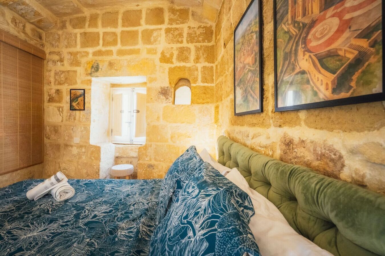 A room with a stone wall, a bed with patterned bedding, a green upholstered headboard, and a small stool.