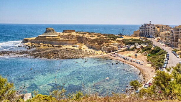 Una località costiera con una penisola rocciosa, acque blu cristalline e una spiaggia con persone e barche.